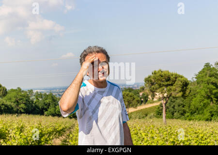 Bel uomo di mezza età con sale pepe capelli vestito con maglietta sportiva è tenendo la sua testa in campi coltivati della campagna italiana: egli sembra avere mal di testa Foto Stock