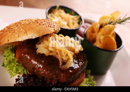 Deliziosa succosa hamburger di manzo con cipolle caramellate e insalata servita con fette di patate su una piastra bianca su di un tavolo di legno. Foto Stock