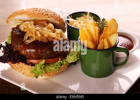 Deliziosa succosa hamburger di manzo con cipolle caramellate e insalata servita con fette di patate su una piastra bianca su di un tavolo di legno. Foto Stock