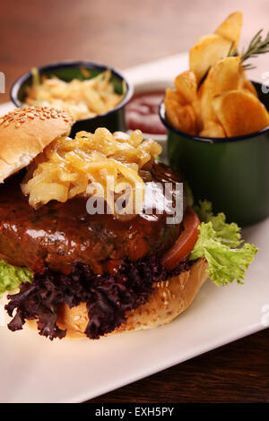 Deliziosa succosa hamburger di manzo con cipolle caramellate e insalata servita con fette di patate su una piastra bianca su di un tavolo di legno. Foto Stock