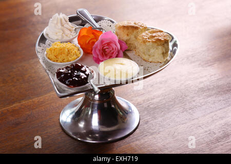 Burro di deliziosi scones su un cuore di argento vassoio sagomato servito con rose crema al formaggio e marmellata conserva su di un tavolo di legno. Foto Stock