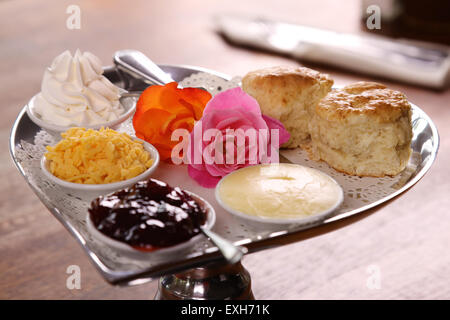 Burro di deliziosi scones su un cuore di argento vassoio sagomato servito con rose crema al formaggio e marmellata conserva su di un tavolo di legno. Foto Stock