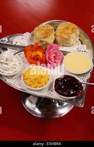 Burro di deliziosi scones su un cuore di argento vassoio sagomato servito con rose crema al formaggio e marmellata conserva su un rosso tavolo in legno. Foto Stock