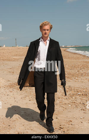 Uomo in un lungo mantello nero in possesso di una pistola e di camminare sulla spiaggia. Foto Stock