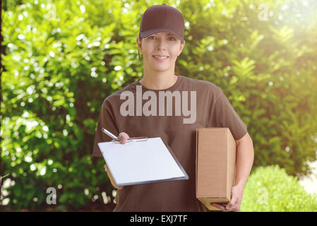 Sorridente attraente corriere femmina in un cappuccio consegnare un pacco alla porta di casa tenendo fuori un blocco appunti per la firma di un Foto Stock
