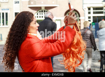 DORDRECHT, Paesi Bassi - 27 Aprile 2015: donna attraente prendendo foto con il telefono cellulare vestito in arancione con lunghi capelli scuri c Foto Stock