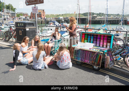 Un commerciante di strada vendita trecce di capelli e intreccio di giovani ragazze con il porto a Padstow Cornwall Regno Unito Foto Stock