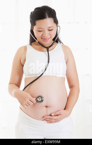 Donna incinta tenendo uno stetoscopio sull'addome con sorriso, Foto Stock