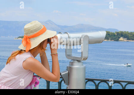 Ragazza giovane guardando attraverso il binocolo pubblica al mare indossando cappello di paglia e abito rosa Foto Stock