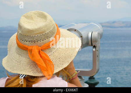 Ragazza giovane guardando attraverso il binocolo pubblica al mare indossando cappello di paglia e abito rosa Foto Stock