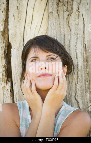 Donna appoggiata contro il tronco di albero con la faccia nelle mani, cercando dreamily Foto Stock