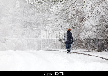 Una donna che cammina lungo una coperta di neve strada suburbana attraverso una tormenta di neve. Utilizzando la strada per guadagnare qualche presa come il sentiero è congelata e scivolose Foto Stock