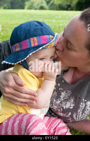 Baby girl succhiare il suo pollice e di essere confortati da sua madre nel giardino Foto Stock