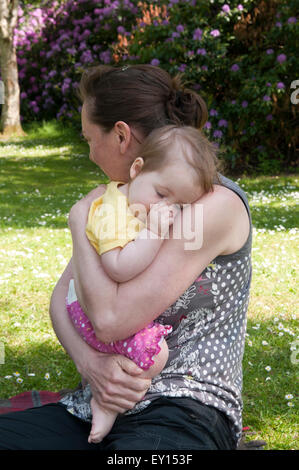 Baby girl succhiare il suo pollice e di essere confortati da sua madre nel giardino Foto Stock