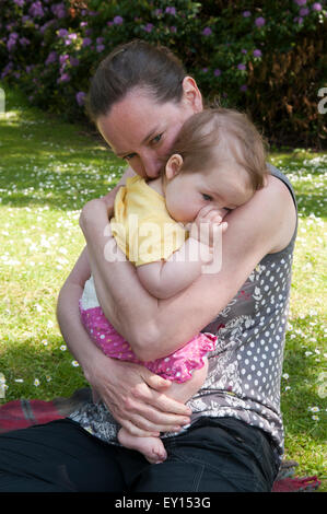 Baby girl succhiare il suo pollice e di essere confortati da sua madre nel giardino Foto Stock