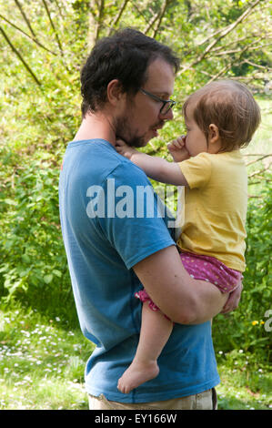 Baby girl succhiare il suo pollice e di essere confortati da suo padre in giardino Foto Stock