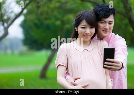 Giovane uomo e donna incinta guardando il telefono cellulare e scattare foto con sorriso, Foto Stock