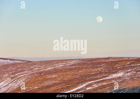 Luna crescente sopra la brughiera con tracce di tardo inverno neve. Belli i colori della luce del sole serale sulla collina. Foto Stock