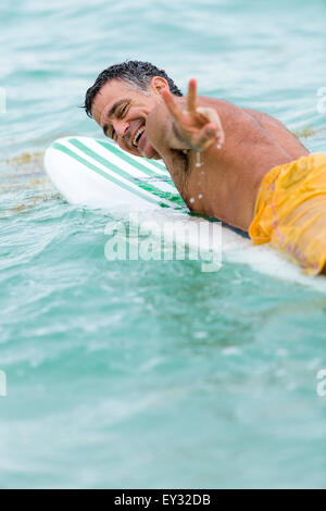 Medium shot di un sano senior che mostra a noi il segno di pace mentre lui è sdraiato sulla sua tavola da surf sull'oceano Foto Stock