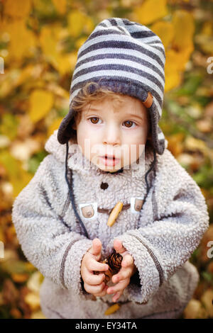 Close-up ritratto di Carino curioso Toddler Boy con grandi occhi in maglia striata Hat cercando in autunno Park. Kid Holding coni. Foto Stock