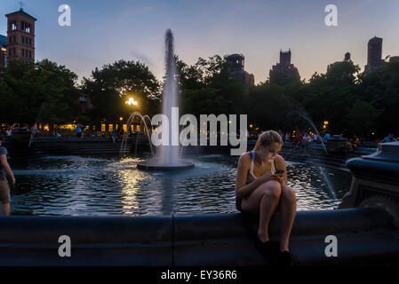 New York, NY - 20 Luglio 2015 - Una donna si siede presso la fontana ed esamina il suo telefono cellulare in una serata estiva in Washington Square Park Stacy Rosenstock Walsh/ALamy Foto Stock