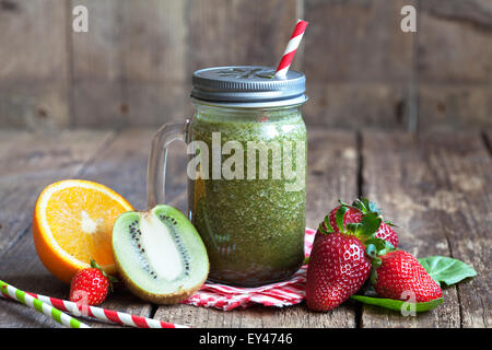 Un sano frullato verde realizzato da spinaci, kiwi, fragole e arance in un vaso con paglia rosso su una tavola di legno Foto Stock