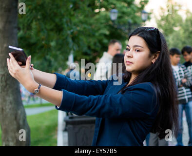 Giovane donna con capelli scuri si prepara a prendere un selfie con il suo smartphone a una fontana pubblica display a Ufa in Russia nel luglio 2015 Foto Stock