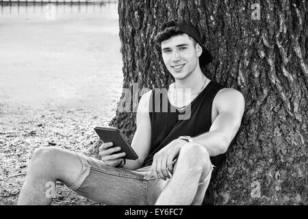 Sorridente giovane uomo bello, la lettura sul lettore di ebook o in possesso di un computer Tablet, rilassante su terreno erboso a Park, aga pendente Foto Stock