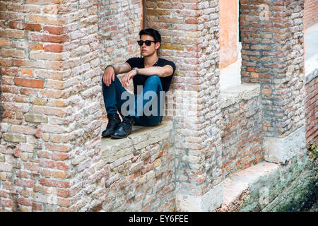 Attraente giovane uomo seduto contro un muro di mattoni, cercando di fronte a se stesso, indossando occhiali da sole Foto Stock