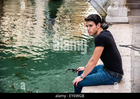 Ritratto di attraente dai capelli scuri giovane uomo seduto Docks accanto a stretto canale a Venezia, Italia Foto Stock