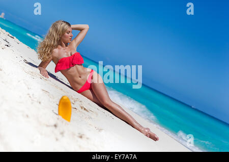 Slim concia di donna con capelli lunghi biondi che giace sulla spiaggia tropicale vicino al Mar dei Caraibi con protezione solare crema in sabbia, o di messa a fuoco Foto Stock