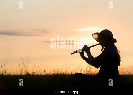 Ragazza adolescente sat in erba giocando un flauto al tramonto. Silhouette Foto Stock