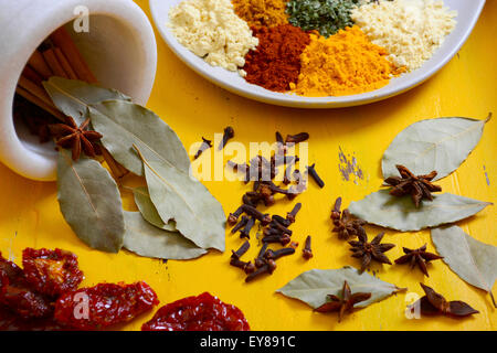 Cucina colorata le spezie e le erbe su un piatto rotondo con bastoncini di cannella, foglie di alloro e anice stellato in un mortaio e pestello su yello Foto Stock