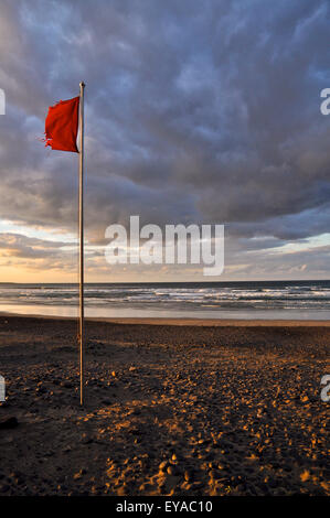 Bandiera logora rossa della spiaggia che si infrangono nel vento, con onde e nuvole al tramonto durante la bassa marea nella spiaggia di Famara (Lanzarote, Isole Canarie, Spagna) Foto Stock