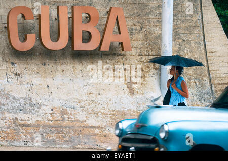 L'Avana, Cuba - Giugno 12, 2011: Vintage americano auto passa pedonale cubano camminando sul marciapiede sotto un grande segno di Cuba. Foto Stock