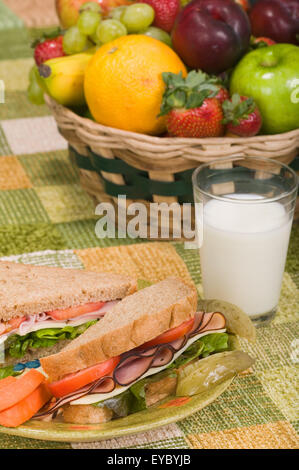 Prosciutto su pane di segale panino con lattuga, pomodoro e formaggio, salamoia e i bastoncini di carote, con una cesta di frutta in background Foto Stock