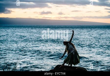 Stagliano donna hula sulle rocce costiere indossando il costume tradizionale al tramonto, Maui, Hawaii, STATI UNITI D'AMERICA Foto Stock