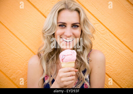 Una giovane donna lambisce un cono gelato mentre in piedi di fronte a un arancio brillante parete. Foto Stock