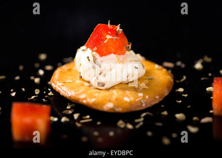 Un cracker con crema di formaggio e peperone rosso su sfondo nero Foto Stock