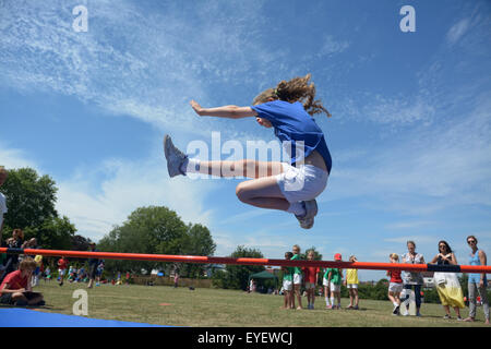 Giovani schoolgirl in una maglietta blu ad alta salta durante la giornata dello sport come compagni di classe e le mamme stanno a guardare in background Foto Stock