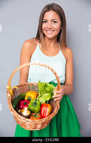 Ritratto di un sorridente giovane donna cestello di contenimento con le verdure sopra uno sfondo grigio Foto Stock