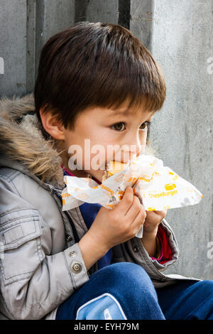 Ragazzo seduto all'aperto nella piccola alcova sul marciapiede, tenendo un McDonald's Cheeseburger in entrambe le mani e mordere in esso. Bambino caucasico, 5-7 anni. Foto Stock