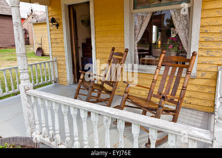 Una coppia di vecchi in legno Sedie a dondolo sedersi sul legno verniciato portico di un giallo sbiadito country house. Foto Stock