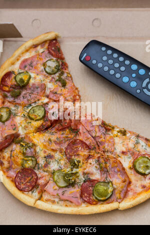 Pezzi di pizza con salsicce e bacon in scatola di cartone con il telecomando del televisore Foto Stock