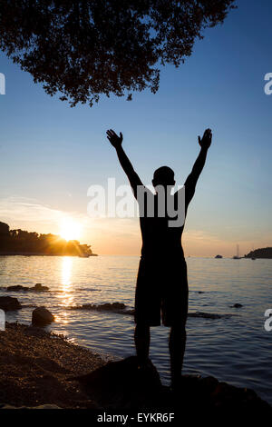 Un uomo in piedi su una roccia sulla costa rivolta verso il mare aperto e salutando il sole con i bracci sollevati a Rovigno Croazia. Foto Stock