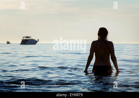 Una vista posteriore di una donna in piedi su un poco profondo sulla costa rivolta verso le barche a vela in mare aperto a Rovigno Croazia. Foto Stock