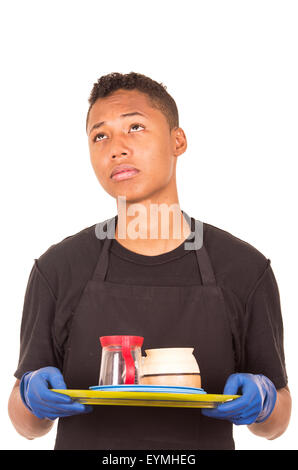 Ispanico giovane uomo blu da indossare guanti di pulizia che porta il vassoio con tazze e fantasticando espressione facciale Foto Stock