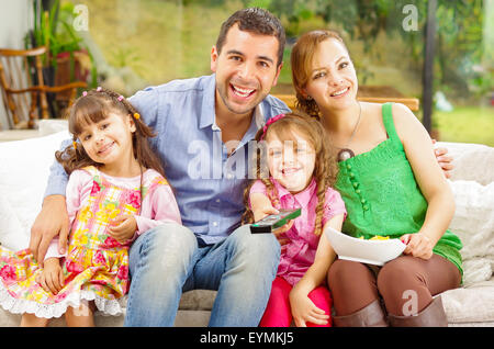 Ritratto di famiglia del padre, madre e due figlie seduti insieme in divano sorridente verso la telecamera che abbraccia ogni altro Foto Stock