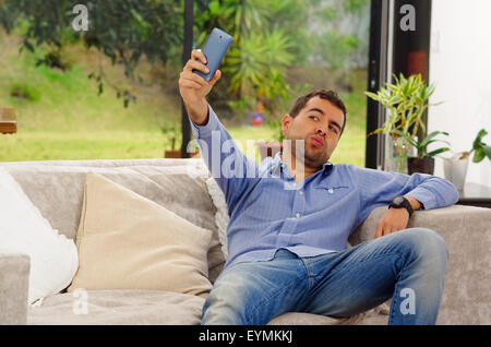 Uomo ispanico indossano jeans e maglietta blu seduta in divano in posa per selfie rendendo le labbra bacio appoggiato all'indietro Foto Stock
