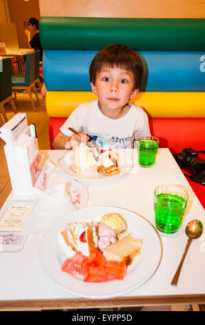 Bambino caucasico, boy, 7-8 anno, seduti al ristorante tavolo con bicchiere di bibita verde e la scorpacciata di torta tenendo cucchiaio, guardando il visualizzatore. Foto Stock